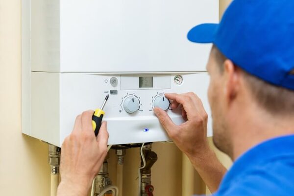 An engineer in a blue cap adjusts the dials of a wall-mounted white combi gas boiler with a screwdriver