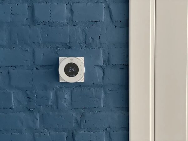 A round white smart room thermostat mounted on a blue painted brick wall next to a white door frame, displaying 24.5 degrees