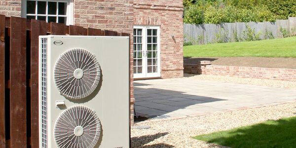 A Grant air source heat pump with two fans installed outside a brick house next to a wooden fence and paved patio