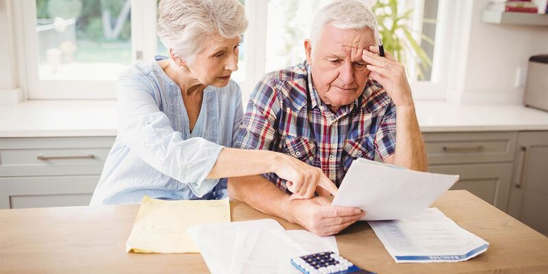 An older couple at a kitchen table looking worriedly at household bills and paperwork with a calculator