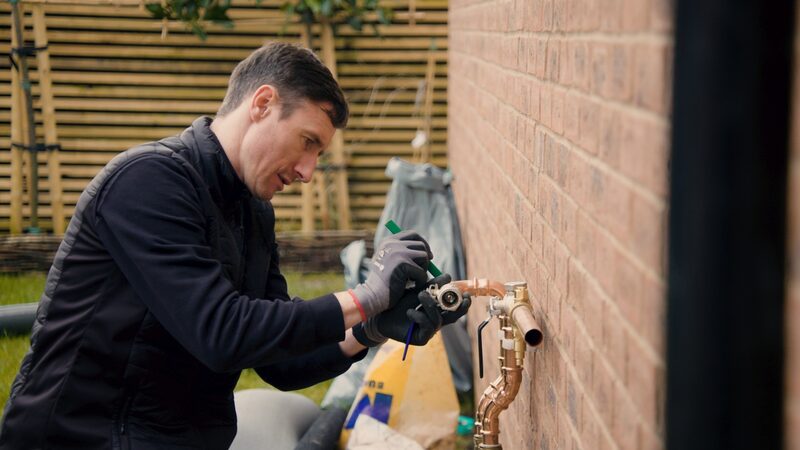 An installer in gloves working on copper pipework and valves mounted on an outside brick wall of a house