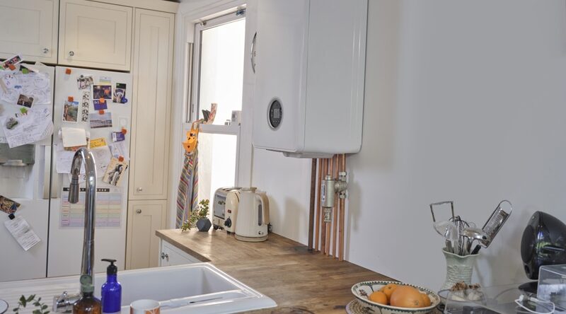 A white wall-mounted combi boiler installed on a kitchen wall above copper pipework, next to a worktop with a kettle and toaster