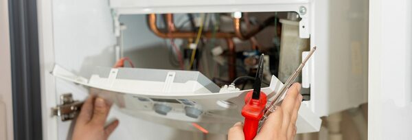 A person repairing a wall-mounted gas boiler with its front panel open, holding a screwdriver and multimeter probes among visible wires and pipes