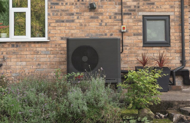 A dark grey air source heat pump unit installed on the exterior brick wall of a house beside a window, surrounded by garden plants