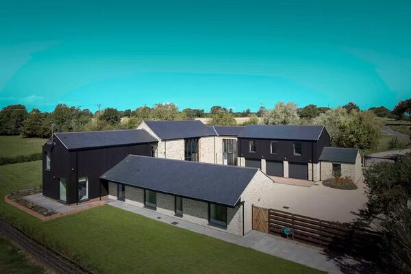 A large modern detached stone-and-dark-clad house with dark solar tile roofs, surrounded by lawns and trees under a blue sky