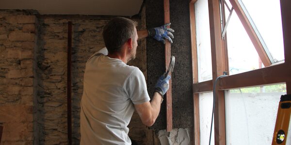 A man fitting wood-fibre insulation around a window reveal during a whole-house retrofit in a stone-walled room