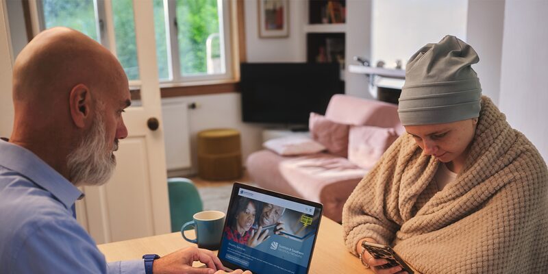 A man and a woman in a living room looking at a laptop showing the Scottish and Southern Electricity Networks website while she uses a phone
