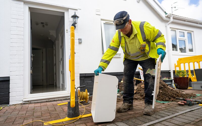 A gas engineer in high-visibility jacket and safety goggles installing a white meter box outside a house during a gas connection works