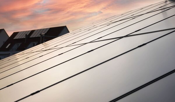 A close-up of solar panels mounted on a roof at sunset, with a neighbouring house with solar panels in the background