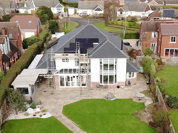 Aerial view of a large detached house with solar panels being installed on its roof, surrounded by scaffolding and neighbouring homes