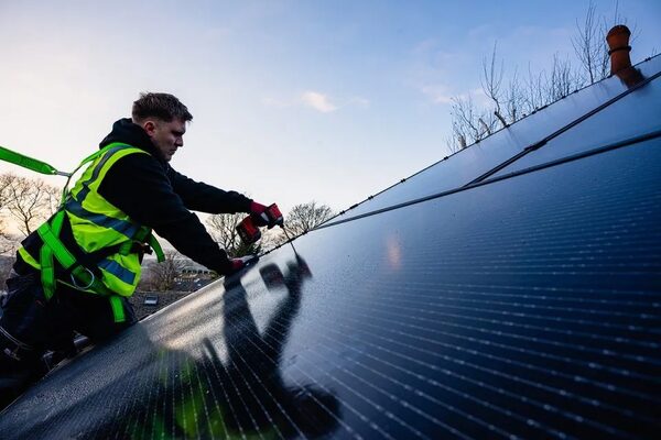 An installer in a high-visibility vest and safety harness fixing a solar panel to a roof with a power drill
