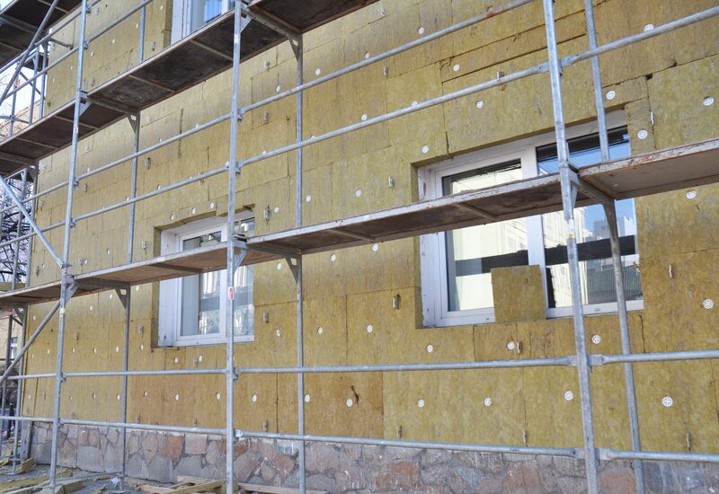 A building facade covered in yellow mineral wool external wall insulation boards behind scaffolding, with windows cut out