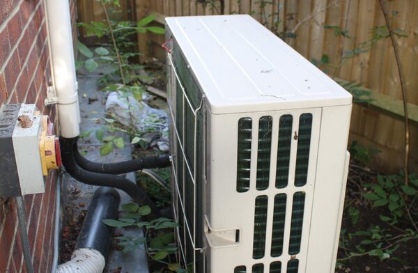 An outdoor air source heat pump unit with grille panels installed beside a brick house wall in a garden