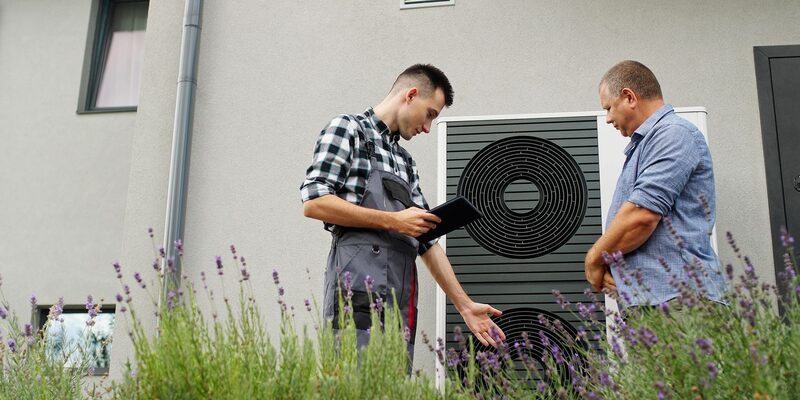 An installer with a tablet showing a homeowner an air source heat pump unit outside a house