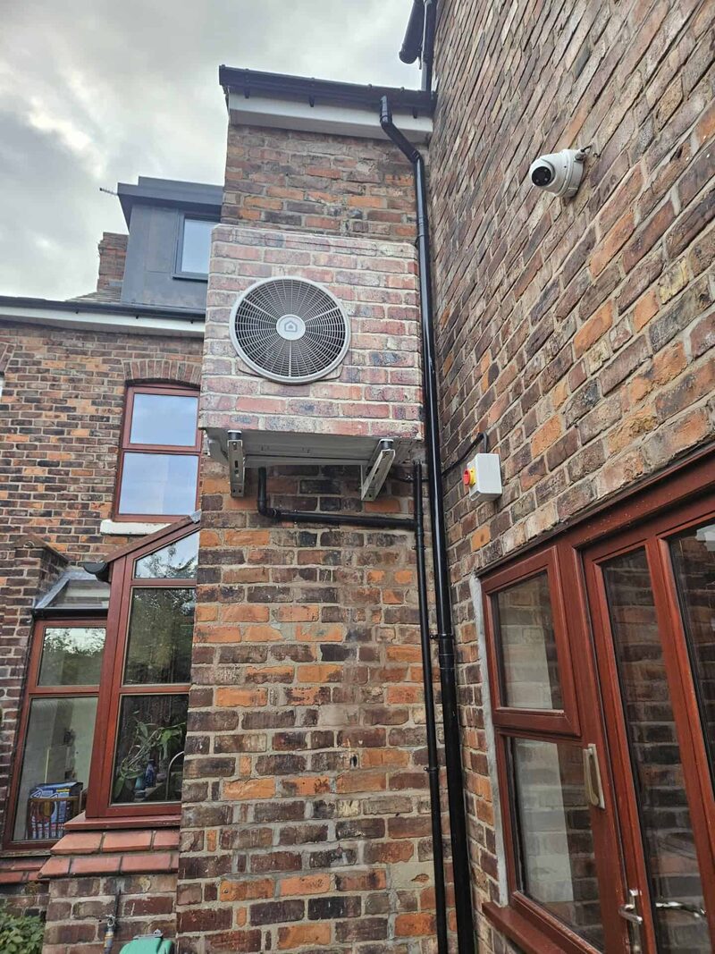An air source heat pump unit mounted on the brick wall of a Victorian terraced house, with a security camera and downpipe nearby.