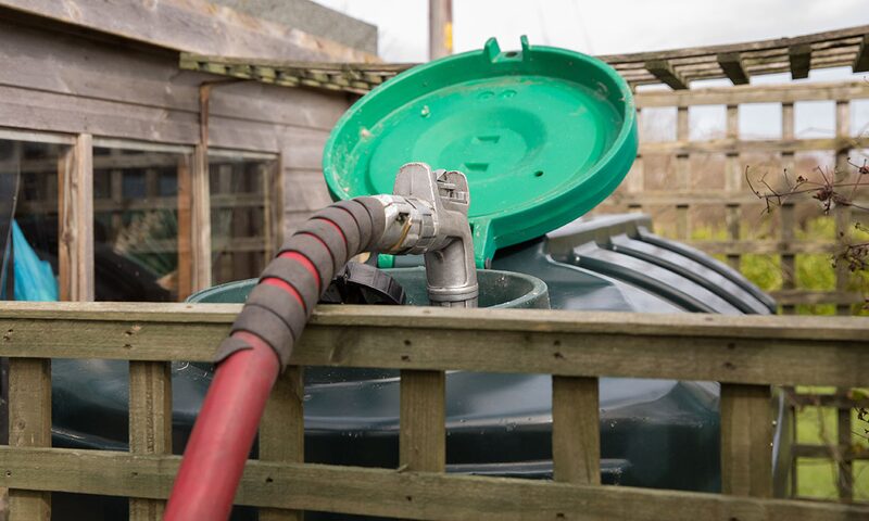 A red delivery hose filling a green domestic heating oil tank behind a wooden fence