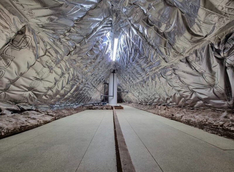 A loft interior lined with reflective foil multifoil insulation draped over the rafters, with boarded floor and a water tank