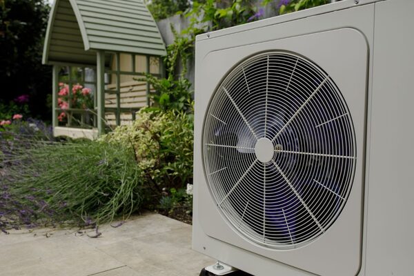 A large air source heat pump outdoor unit with a fan grille installed in a garden beside a paved patio, with a green arbour and planting behind