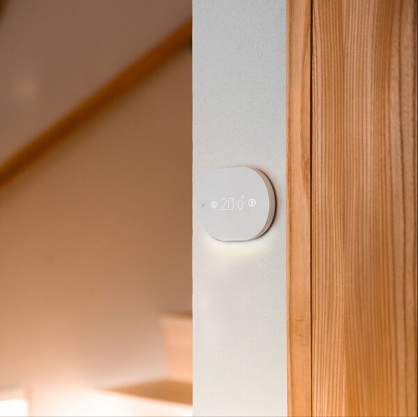 A white round smart thermostat mounted on a wall next to a wooden door frame, displaying a set temperature of 20.0 degrees