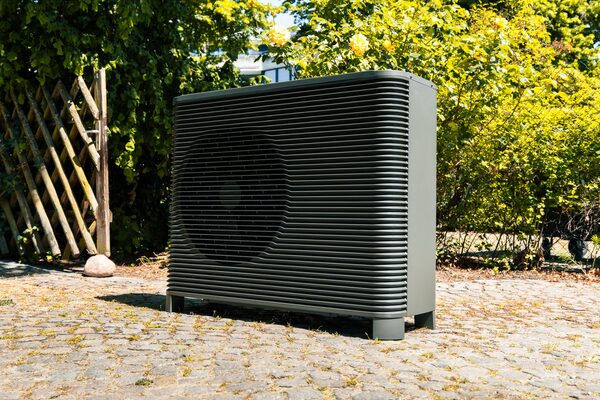 A grey outdoor air source heat pump unit standing on a cobbled surface in a garden