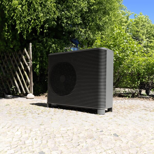 A grey outdoor air source heat pump unit standing on a cobbled driveway in a garden