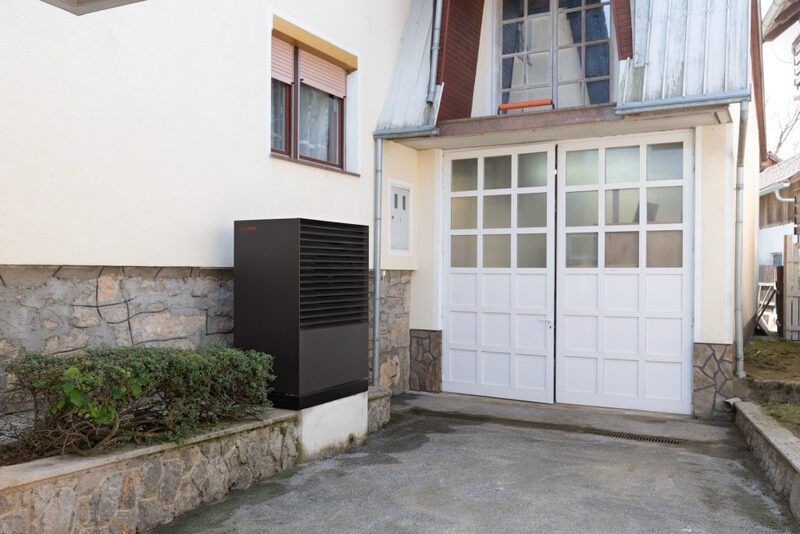 An outdoor air source heat pump unit installed against the exterior wall of a house beside garage doors