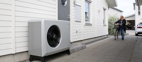 An air-to-water heat pump unit installed outside a white timber-clad house with a woman walking past with a bicycle