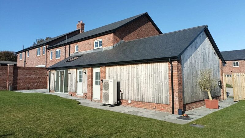 An air source heat pump unit installed on the exterior wall of a modern brick and timber-clad house with a lawn in front