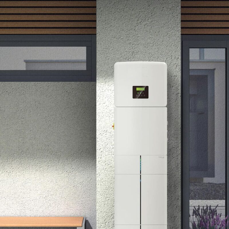 A white wall-mounted home battery storage unit with a small display, installed on an exterior house wall