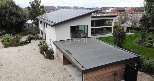 A modern flat-roofed house with solar panels on the sloped roof section, surrounded by garden and new-build homes