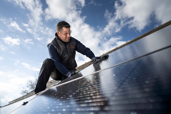 An installer in a gilet and safety glasses fitting a solar panel on a roof under a blue sky