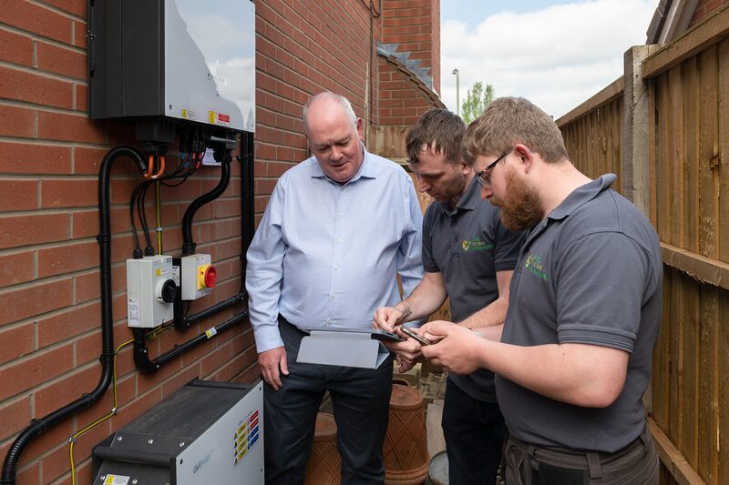 Two installers and a homeowner reviewing paperwork beside a wall-mounted solar inverter and battery unit on a brick house wall