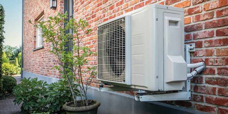 A white air source heat pump unit mounted on the exterior brick wall of a house, next to a potted plant