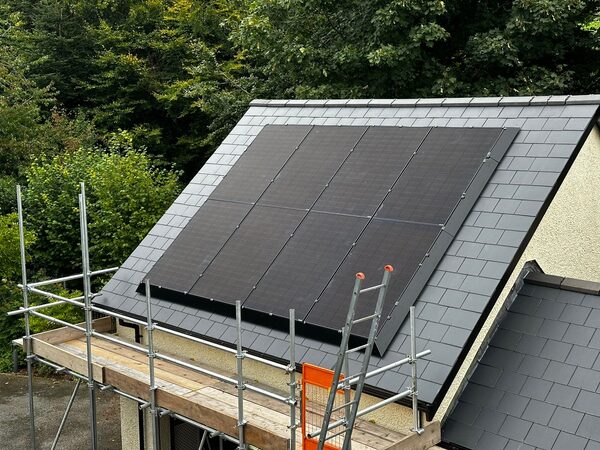 A slate-roofed house with black solar panels installed on the roof, surrounded by scaffolding during installation