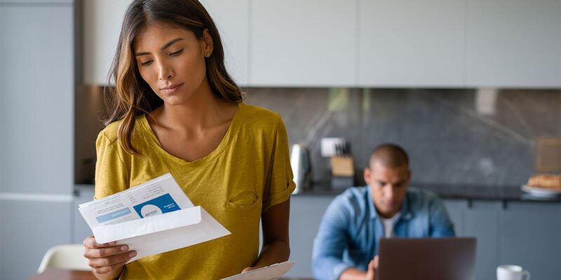 A woman reading a letter from an envelope in a kitchen while a man works on a laptop behind her