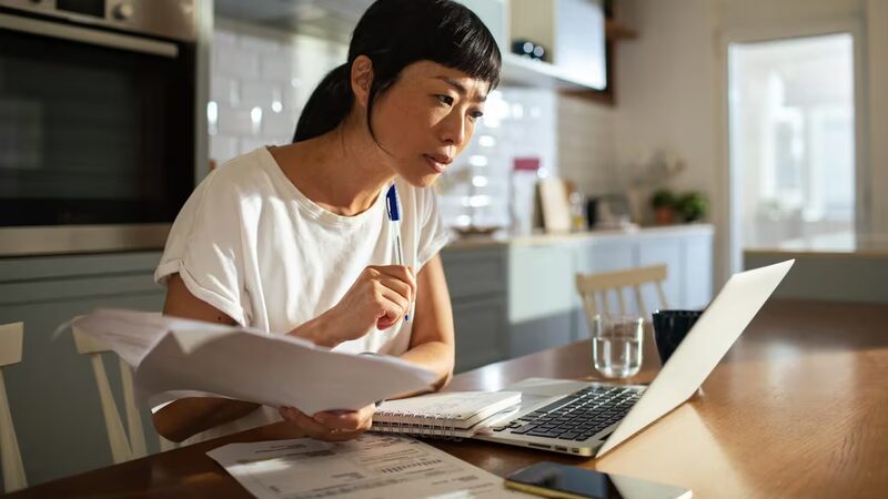 A woman at a kitchen table reviews paperwork while looking at a laptop, with bills and a notebook in front of her