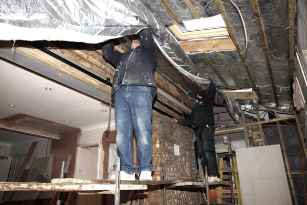 Two workers on scaffolding fitting foil insulation under the exposed rafters of a roof during a home renovation