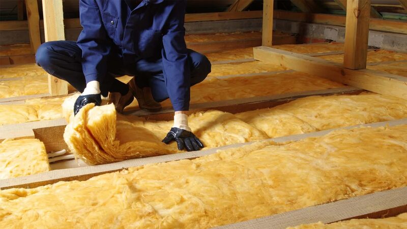 A worker in blue overalls and gloves laying yellow mineral wool insulation between joists in a loft