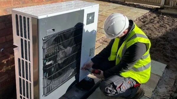An installer in a hard hat and hi-vis vest working on an outdoor air source heat pump unit outside a brick house