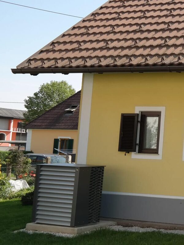 An outdoor heat pump unit installed on a concrete base beside a yellow house with a tiled roof
