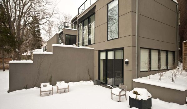 A modern grey-rendered house with large glazed doors and windows, seen from a snow-covered garden in winter