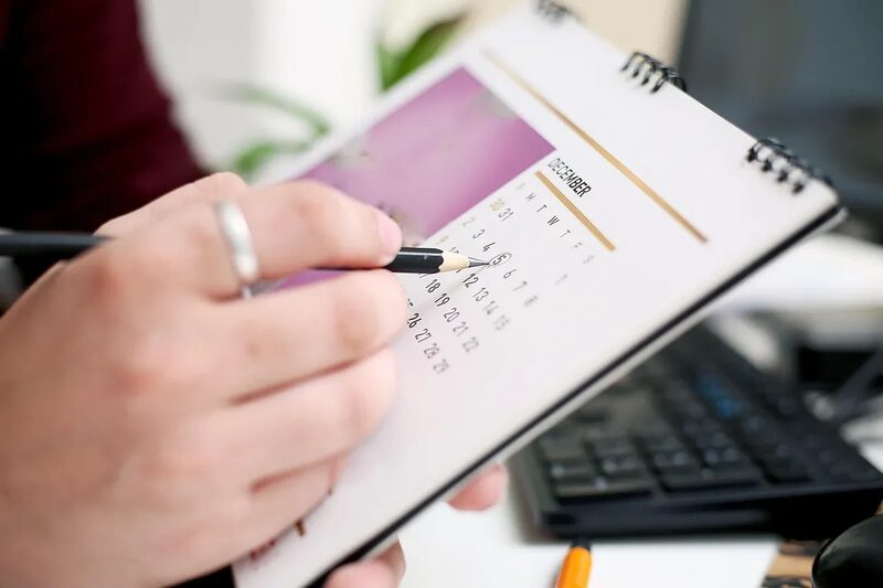 A person's hand marking a date on a desk calendar with a pencil next to a keyboard