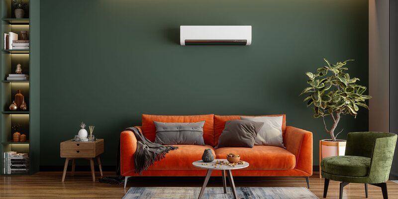 A white wall-mounted air-to-air heat pump unit above an orange sofa in a green living room