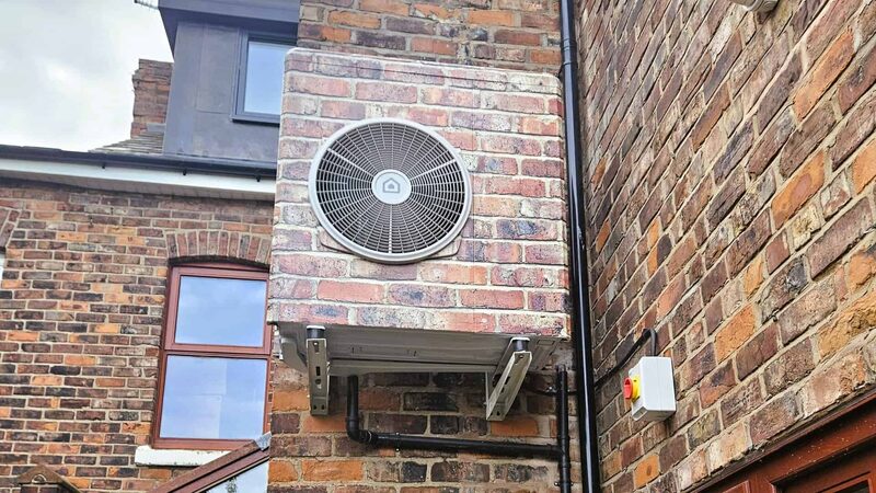An air source heat pump outdoor unit mounted on a wall bracket against the brick exterior of a house