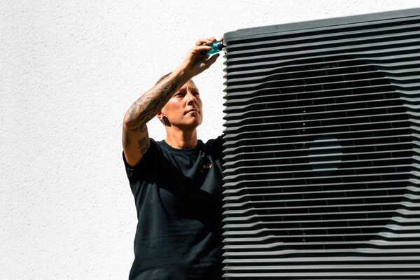 A technician working on an outdoor air source heat pump unit mounted on a white wall