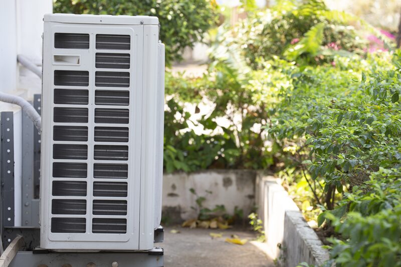 An outdoor heat pump unit mounted on a wall beside a garden path with green shrubs