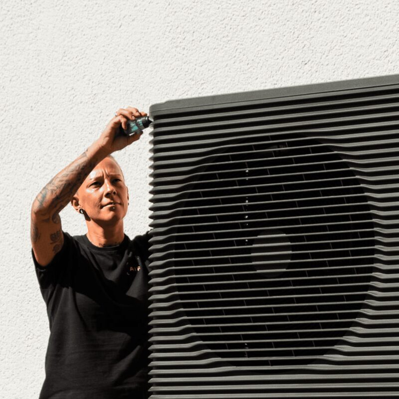 An installer working on an outdoor air source heat pump unit against a white wall