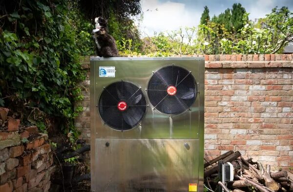 A large stainless steel air source heat pump unit with two fans installed outdoors in a garden next to a brick wall, with a cat sitting on top