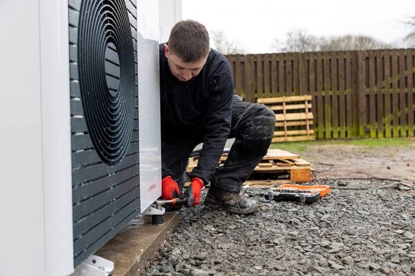 An engineer in work clothes and red gloves installing an outdoor air source heat pump unit on a gravel surface