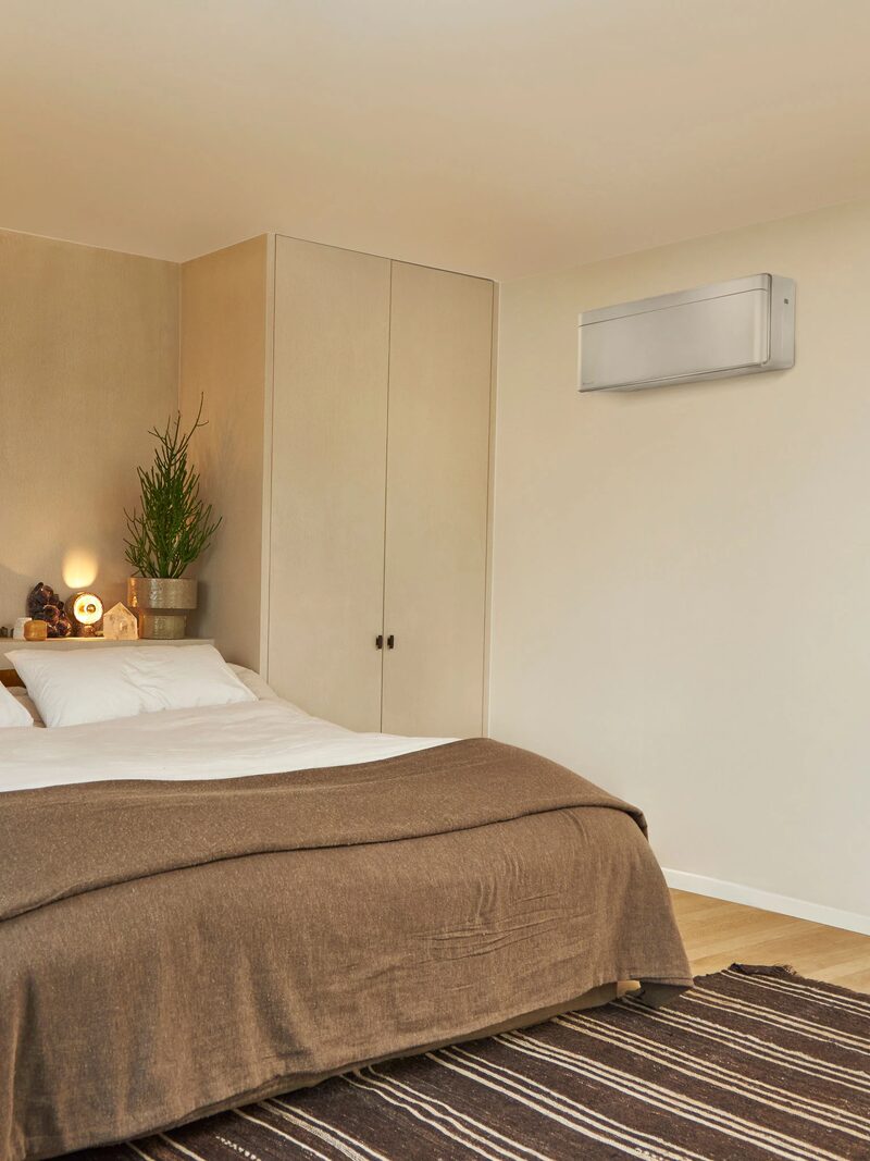 A wall-mounted air-to-air heat pump indoor unit installed high on the wall of a cosy bedroom with a large bed